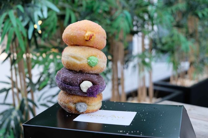 Stack of four sugar-dusted filled donuts with colorful cream and jam fillings on a black pastry box, set on a table with blurred bamboo plants in the outdoor cafe background.