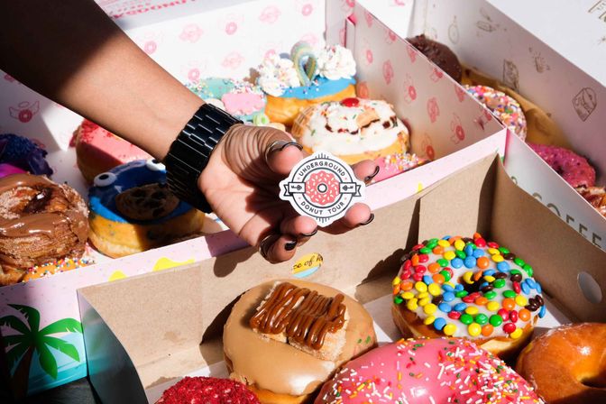 Close-up of a hand holding a donut-themed enamel pin above open boxes of vibrant gourmet donuts—sprinkles, colorful candy-coated chocolates, pink and caramel glazes.