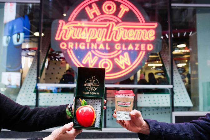 Two hands present a shiny apple-shaped glazed donut in a green box and a takeaway coffee cup beneath a bright neon "Hot Now / Original Glazed" sign outside a Times Square doughnut shop