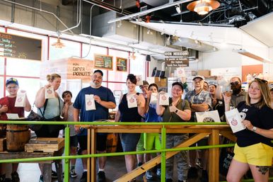 Group of smiling customers inside a bright local cider-donut shop at an indoor farm market, each holding a small paper bag of treats.