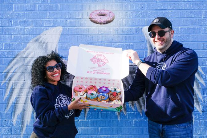 Two smiling people in navy hoodies and sunglasses hold open a box of colorful, decorated donuts in front of a blue brick wall featuring a painted donut mural.