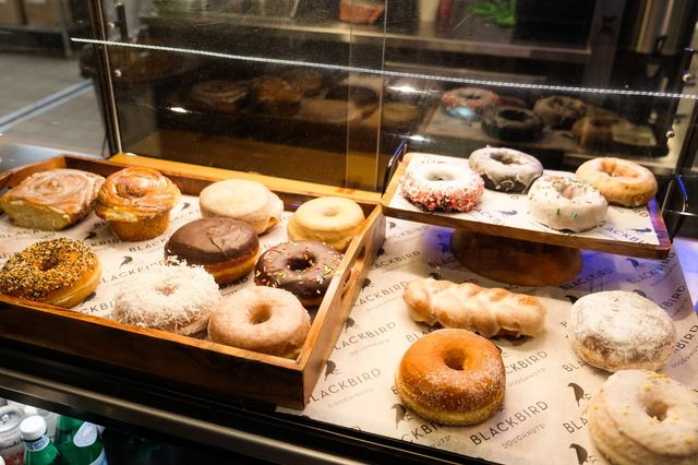 Assorted donuts and pastries (glazed, chocolate, sprinkled, coconut) arranged on wooden trays inside a glass bakery display case