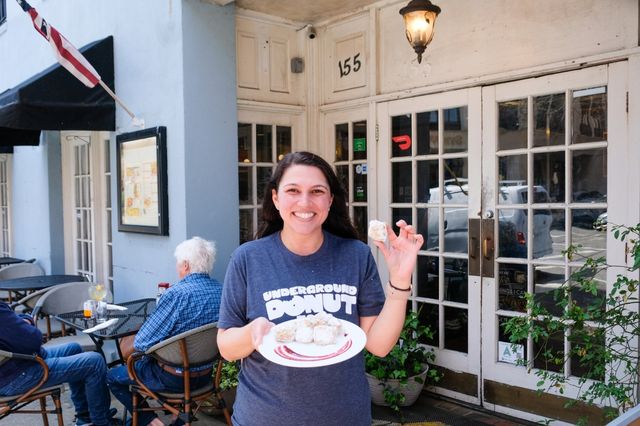 Smiling woman holding a plate of powdered donut bites and one donut in hand outside a sidewalk café with glass‑paned doors and outdoor patio seating