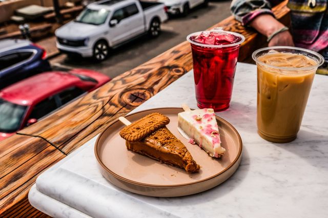 Outdoor cafe marble table with two popsicle-style dessert slices (caramel cookie and white berry) on a plate, a red iced tea and an iced coffee in plastic cups, wood balcony railing and parked cars in the background.