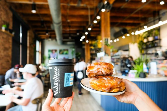 Takeaway coffee cup and two glossy glazed donuts on a small plate held up inside a busy modern industrial café with wooden beams, exposed ductwork, hanging lights and people working at tables.