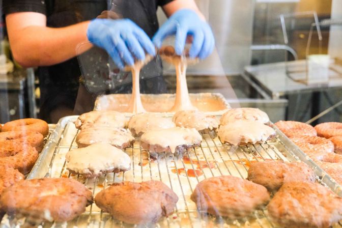 Gloved hands pouring creamy glaze over freshly fried donuts on a cooling rack behind a bakery display window.