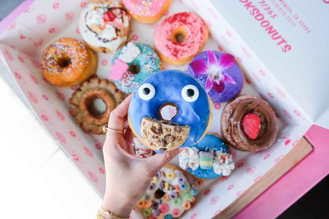 Top-down view of a hand holding a blue-frosted donut with candy eyes and a cookie “mouth,” above a box of colorful decorated gourmet donuts with sprinkles, fruit and cereal toppings, playful and bright.