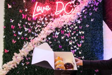 Open box of colorful gourmet donuts held by a hand in front of a butterfly-covered floral wall with a pink neon sign reading Love DC in Washington, D.C.