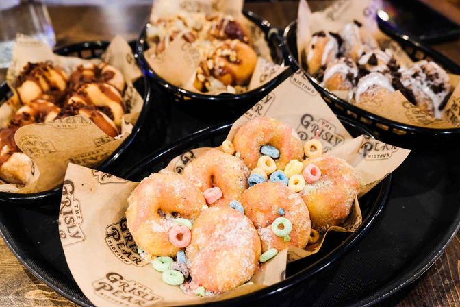 Close-up of assorted mini donuts in black baskets; foreground shows sugar-coated mini donuts sprinkled with colorful cereal rings, with chocolate-drizzled donut bites visible in the background.