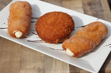 Two sugar-coated, cream-filled fried pastries flanking a golden, crispy breaded donut on a white rectangular plate over a wooden table.
