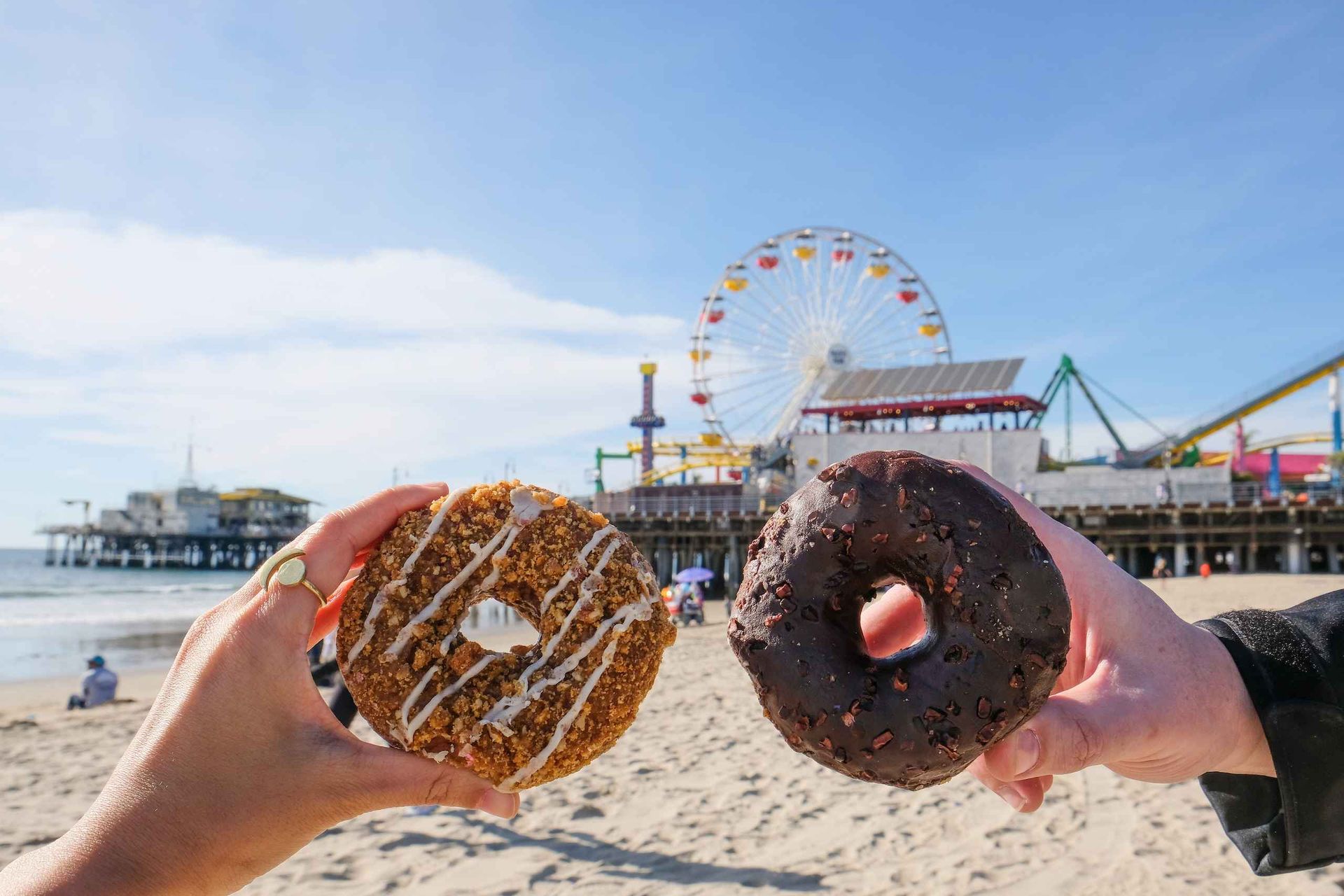 Two hands holding donuts—crumb-topped with white drizzle and chocolate-glazed—on a sunny sandy beach with a Ferris wheel and oceanfront amusement pier in the background.