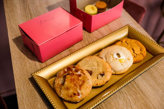 Four assorted gourmet cookies, including a flower-topped lemon cookie, arranged on a gold decorative tray beside pink bakery boxes and a small box of macarons on a wooden table