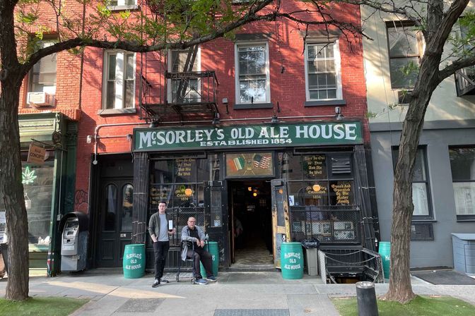 Historic East Village brick pub exterior with green Old Ale House sign reading Established 1854, open doorway and two people on the sidewalk under street trees
