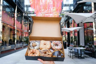 Hand holding an open box of six assorted gourmet donuts in front of glass storefronts and a pink ribbon canopy at an outdoor urban shopping plaza