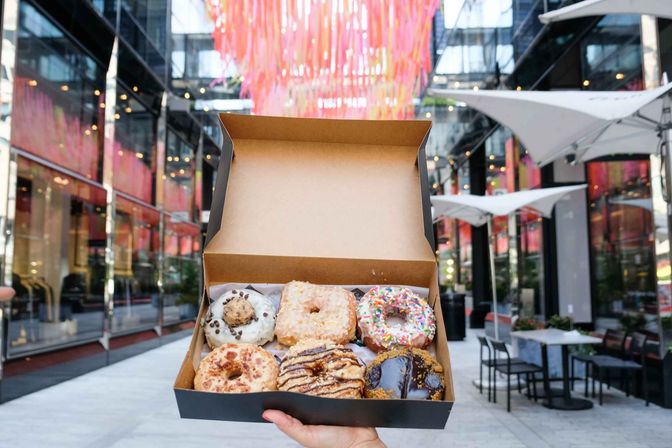 Hand holding an open box of six assorted gourmet donuts in front of glass storefronts and a pink ribbon canopy at an outdoor urban shopping plaza
