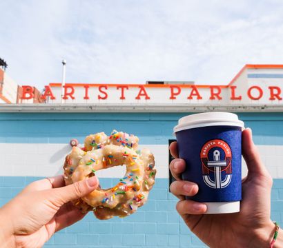 Hands holding a sprinkled glazed donut and a takeaway coffee cup against a bright blue cafe exterior with orange signage