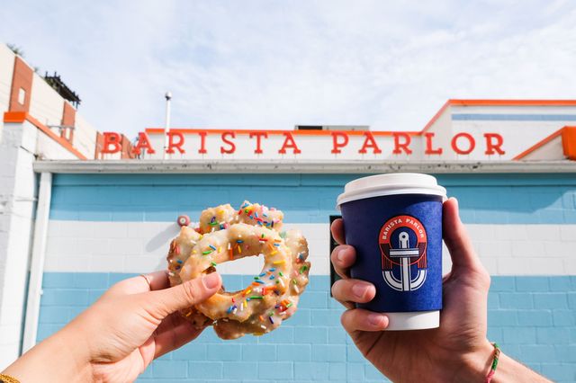 Hands holding a sprinkled glazed donut and a takeaway coffee cup against a bright blue cafe exterior with orange signage