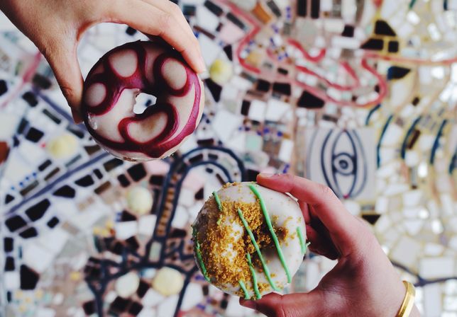 Two colorful donuts held by hands above a vibrant mosaic tile surface — white-glazed with green drizzle and golden crumbs, and white-glazed with purple swirl.