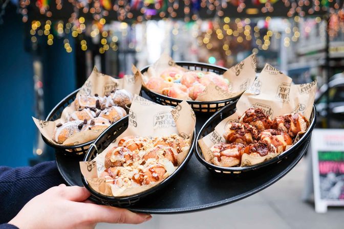 Hands holding a tray of four baskets of mini doughnuts with powdered sugar, chocolate drizzle, crushed nuts and colorful cereal, served as street food on a city sidewalk under festive bokeh lights.