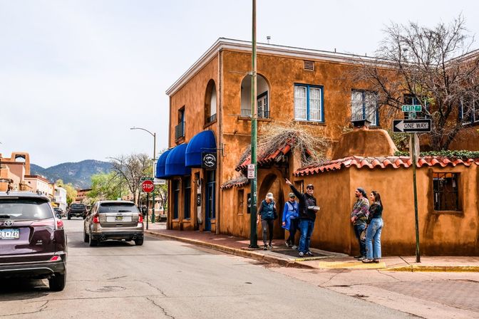 Santa Fe‑style adobe street corner with blue awnings, pedestrians chatting on the sidewalk, parked cars, one‑way and stop signs, and distant foothills under an overcast sky