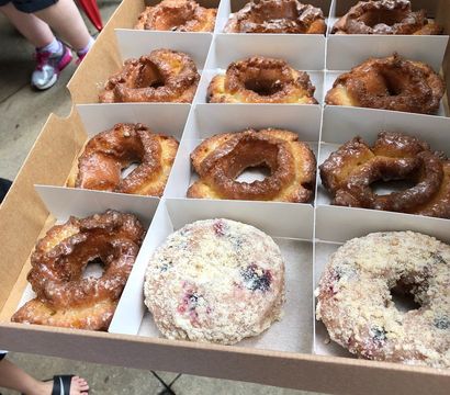 Bakery takeout box of fresh, crispy glazed old-fashioned donuts with two crumb-topped berry donuts, held outdoors on a sidewalk.