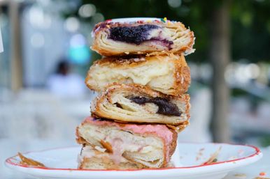 Close-up stack of flaky filled pastries on a plate — blueberry, vanilla custard, chocolate, and strawberry-glazed layers — photographed at an outdoor café.