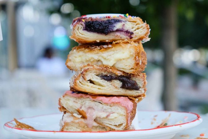 Close-up stack of flaky filled pastries on a plate — blueberry, vanilla custard, chocolate, and strawberry-glazed layers — photographed at an outdoor café.