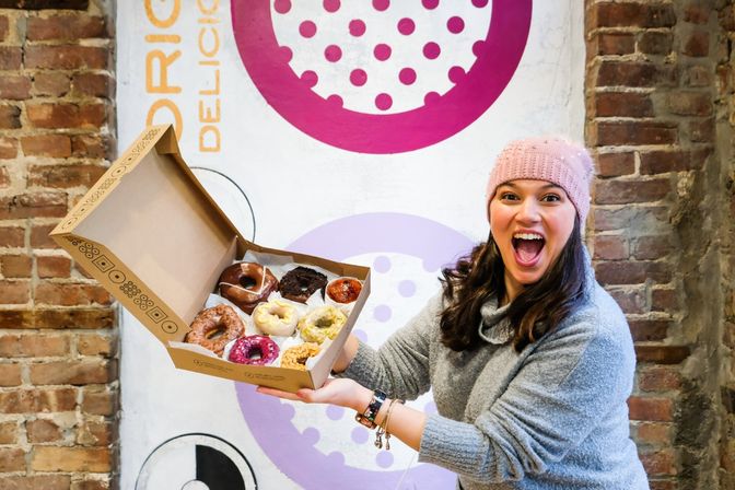 Excited woman in a pink knit hat and gray sweater holding an open box of assorted colorful donuts in front of a brick wall with a large pink donut mural