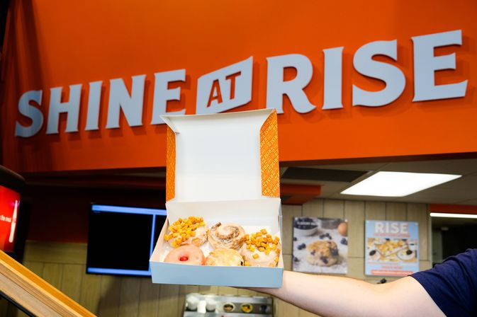 Hand holding an open white box of assorted glazed donuts and cinnamon rolls topped with cereal bites, inside a bright orange breakfast cafe interior with large white wall lettering and menu posters in the background.