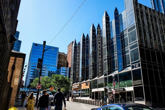 Sunny downtown street scene: pedestrians walk past a reflective glass office building with pointed vertical spires, mirrored skyscrapers and a clear blue sky.