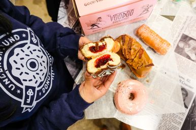 Hands holding a torn-open jelly-filled chocolate-glazed doughnut, surrounded by assorted donuts — pink-frosted sprinkle ring, caramel long john with crunchy topping, and a sugar-coated bar — on a newspaper-lined table next to a pink doughnut box and a person wearing a dark donut-logo sweatshirt.