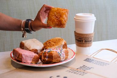 Hand holding a glazed square pastry above a plate of assorted frosted cruffins with sprinkles, next to a takeaway coffee cup on a cafe table