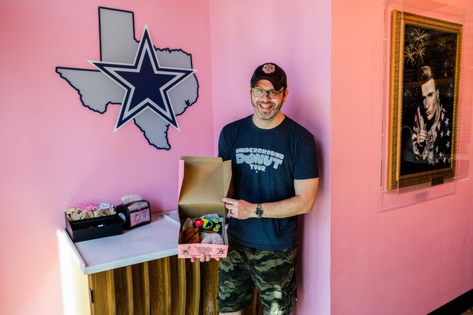 Smiling man in a cap holding an open box of colorful donuts inside a bright pink donut shop with a Texas-shaped wall sign featuring a large star and a framed portrait on the wall.