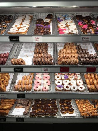 Glass bakery display case with trays of assorted donuts — glazed rings, chocolate-topped, colorful-sprinkled, pink-frosted and cake donuts neatly arranged on metal racks.