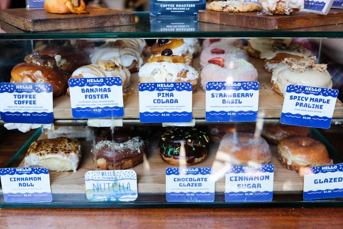 Glass display case at an artisan donut shop showcasing assorted gourmet donuts on wooden trays with blue flavor cards — Toffee Coffee, Bananas Foster, Pina Colada, Strawberry Basil, Spicy Maple Praline, Chocolate Glazed, Cinnamon Sugar — colorful glazes and toppings.