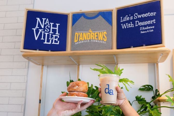 Two sugar doughnuts on a plate and an iced latte held up in a bright Nashville bakery café with leafy plants and wall signage