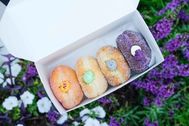 White takeout box holding four sugar-coated filled donuts with colorful cream centers (yellow, green, speckled gray, purple) displayed over vibrant purple and white garden flowers.