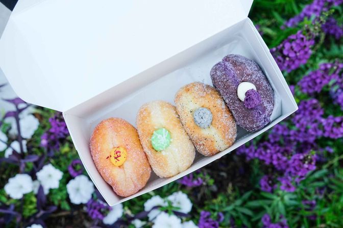 White takeout box holding four sugar-coated filled donuts with colorful cream centers (yellow, green, speckled gray, purple) displayed over vibrant purple and white garden flowers.