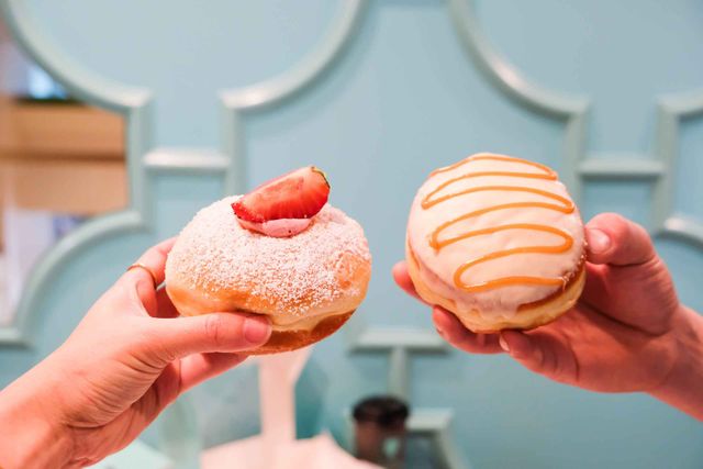 Two gourmet filled donuts held in hands against a pastel blue cafe wall — a powdered sugar donut topped with a strawberry slice and a white-glazed donut with caramel drizzle.