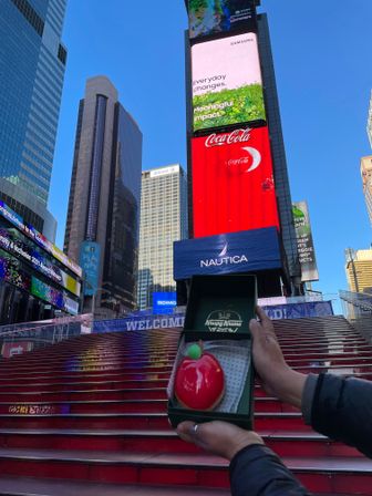 Gleaming red apple-shaped pastry in a green box held over the iconic red stairs beneath towering digital billboards in Times Square, New York City