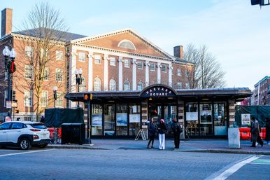 Harvard Square station entrance in Cambridge, MA — curved canopy and sign in front of a historic red‑brick building with white columns; three people chatting on the sidewalk, parked car and bicycles nearby.