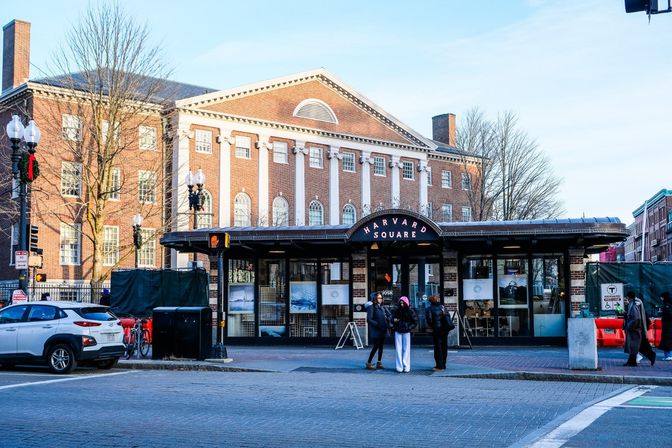 Harvard Square station entrance in Cambridge, MA — curved canopy and sign in front of a historic red‑brick building with white columns; three people chatting on the sidewalk, parked car and bicycles nearby.