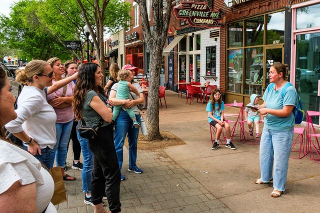 Tour group on a tree-lined downtown sidewalk outside cafes with outdoor seating, a guide speaking to families while kids sit on bright pink chairs — lively urban stroll scene.