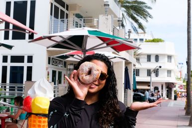Woman with sunglasses playfully holds a powdered-sugar donut over one eye like a monocle on a sunny, palm-lined coastal promenade with an ice cream shop, colorful umbrellas, and outdoor seating.