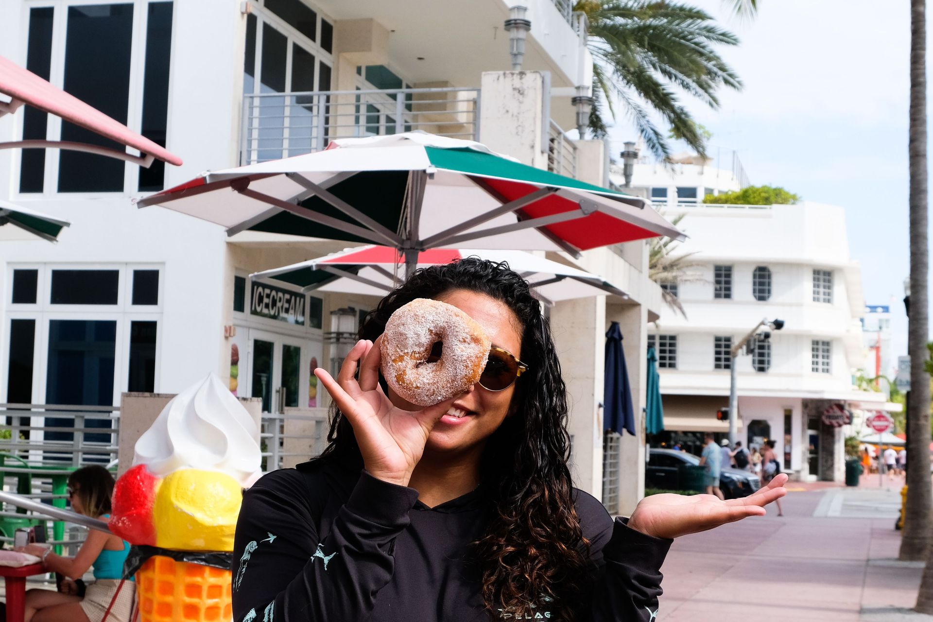 Woman with sunglasses playfully holds a powdered-sugar donut over one eye like a monocle on a sunny, palm-lined coastal promenade with an ice cream shop, colorful umbrellas, and outdoor seating.