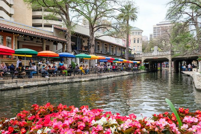 Vibrant outdoor dining along the San Antonio River Walk with colorful umbrellas over riverside tables, pink flowers in the foreground, an arched pedestrian bridge and city buildings in the background.