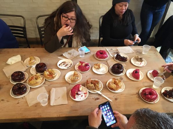 Overhead shot of a wooden cafe table covered with assorted glazed and bright pink-frosted donuts on paper plates while people sample pastries and check phones