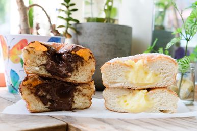 Close-up of two stacked sugar-coated filled doughnuts on a wooden table — gooey chocolate-filled doughnut on the left and creamy vanilla custard-filled doughnut on the right, with potted plants blurred in the background.
