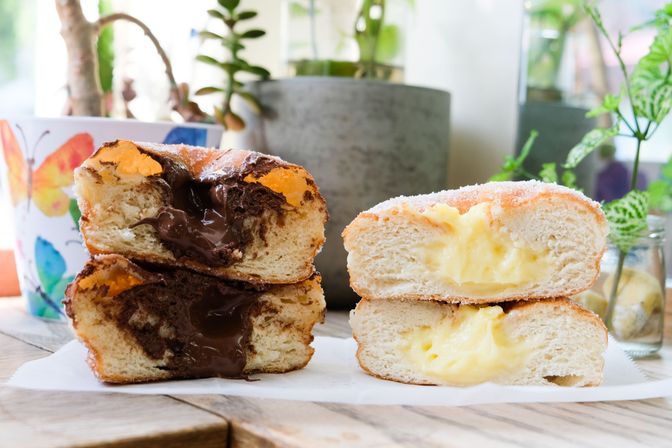 Close-up of two stacked sugar-coated filled doughnuts on a wooden table — gooey chocolate-filled doughnut on the left and creamy vanilla custard-filled doughnut on the right, with potted plants blurred in the background.