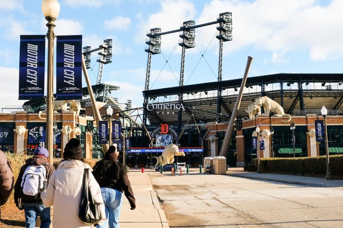 Downtown Detroit baseball stadium entrance with giant feline sculptures, tall floodlight towers, angled metal beams and 'Motor City' banners as pedestrians walk toward the gates on a sunny day.